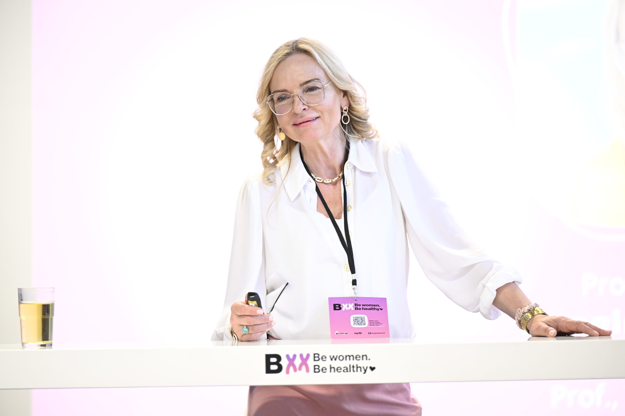 Blonde Frau mit Brille steht lächelnd hinter einem Rednerpult bei einer Veranstaltung. Auf dem Rednerpult steht das Logo ‚BXX Be women. Be healthy.‘ sowie ein Glas mit Getränk.