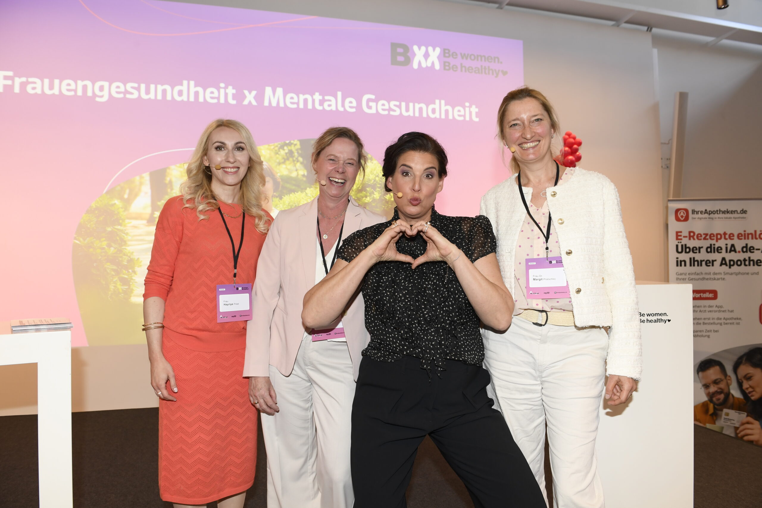 Vier Frauen stehen lächelnd nebeneinander vor einer Präsentationsfolie mit der Aufschrift ‚Frauengesundheit x Mentale Gesundheit‘. Eine Frau in schwarzem Kleid formt mit ihren Händen ein Herz. Alle tragen Namensschilder, die Stimmung ist freundlich und professionell.