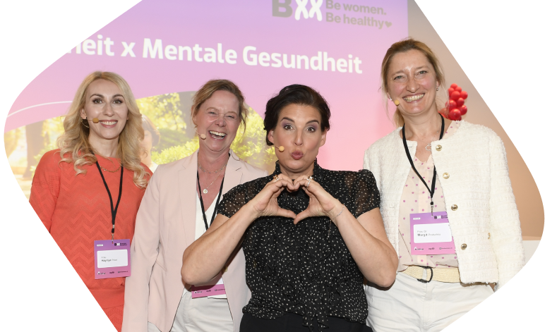 Vier Frauen stehen lächelnd nebeneinander vor einer Präsentationsfolie mit der Aufschrift ‚Frauengesundheit x Mentale Gesundheit‘. Eine Frau in schwarzem Kleid formt mit ihren Händen ein Herz. Alle tragen Namensschilder, die Stimmung ist freundlich und professionell.